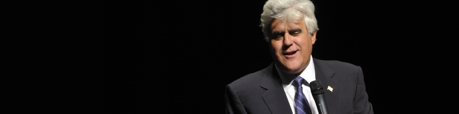 Comedian Jay Leno holding a microphone, standing in front of a black background