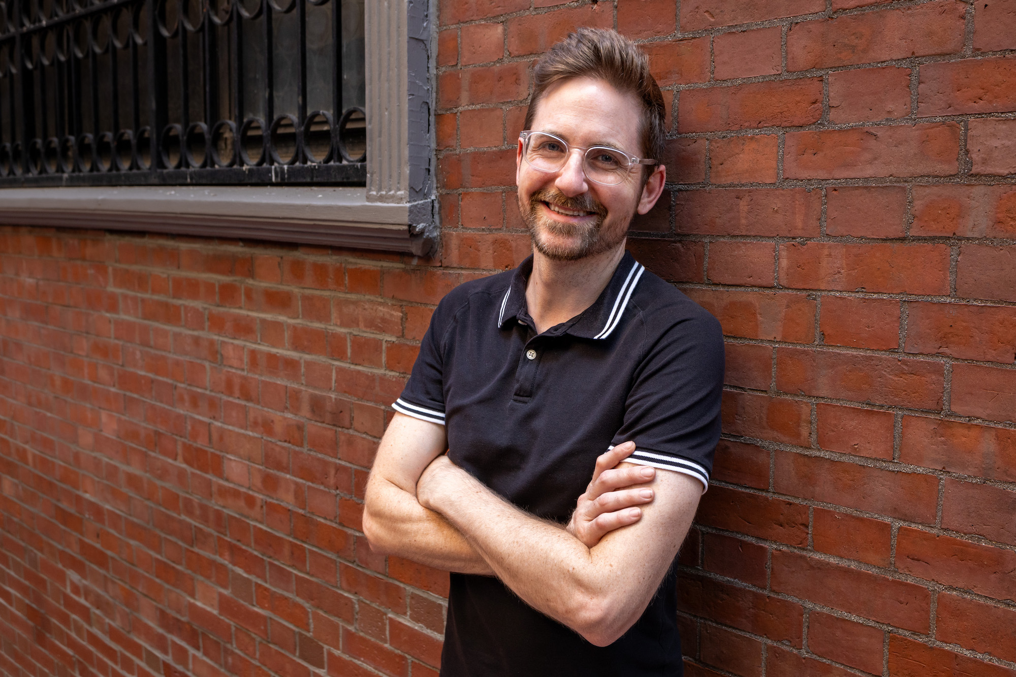 Matthew McMahan wearing a black shirt and glasses, standing in a brick alley